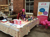 A NETA stall outdoors, with a pull-up banner, pink balloons and goods laid out on a table